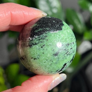 May include: A polished sphere of green zoisite with black inclusions. The gemstone is held between fingers, showcasing its smooth, rounded form and natural color variations. The sphere's surface reflects light, highlighting its texture and the contrast between the green and black.