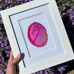 Puede incluir: Una rebanada de ágata rosa enmarcada con bandas blancas y rosas. La ágata está colocada en un passe-partout y un marco blancos.