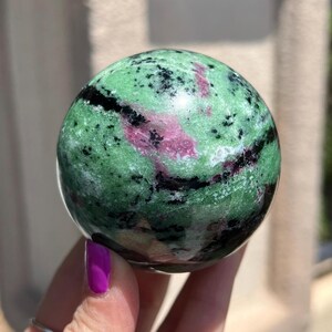May include: Green, pink, and black gemstone sphere. The sphere is polished and has a smooth surface.
