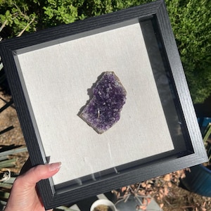 May include: A square black shadow box frame containing a single, irregular piece of deep purple amethyst crystal cluster centered on a light beige fabric backing. The frame features a textured finish and a clear glass front panel.