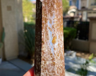 May include: A tall, polished crystal obelisk with a pointed top. The stone features a mix of brown, white, and gold colors. The crystal is held in a hand with blue nail polish, with a blurred outdoor background.