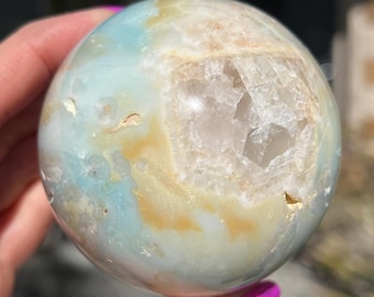 May include: A polished blue, white, and brown agate sphere with a large, clear crystal druzy inside. The sphere is smooth and has a natural, organic look.