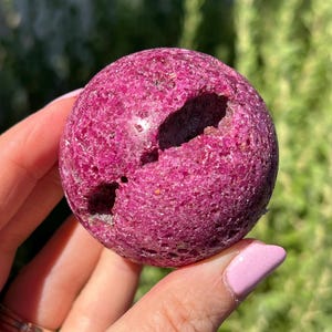 May include: A spherical object with a vibrant magenta, porous, and crystalline surface texture. The sphere features two distinct, irregular, and deep indentations or cavities that reveal a darker, rough interior structure within the stone.
