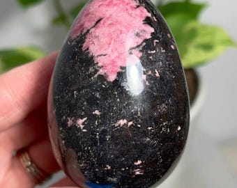 May include: A polished, egg-shaped, pink and black gemstone. The gemstone has a smooth, shiny surface and a swirling pattern of pink and black.
