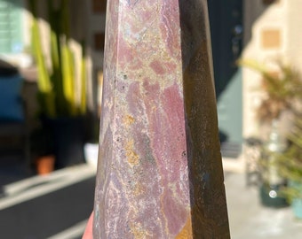 May include: A large, multi-faceted, pink and brown crystal point with a pointed tip. The crystal has a natural, rough texture and is held in a hand.
