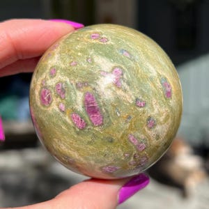 May include: Green and pink sphere with a speckled pattern. The sphere is made of a natural stone and has a smooth, polished surface.
