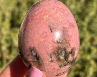 May include: A pink and gray polished Rhodonite egg. The egg is smooth and has a natural, organic shape.