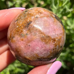 May include: A polished, spherical gemstone with a mix of pink, brown, and black hues. The stone has a smooth, rounded shape and a glossy finish. The gemstone is held in a hand against a backdrop of green foliage.
