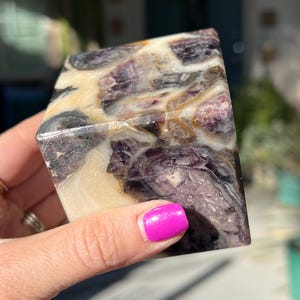 May include: A cube-shaped polished stone with a swirling pattern of purple, white, and brown. The stone has a smooth, polished surface.
