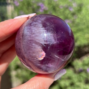 May include: A polished, spherical amethyst crystal ball. The sphere is a deep purple color with white and iridescent inclusions. The crystal reflects light, creating a shimmering effect. The background is a blurred view of green foliage.