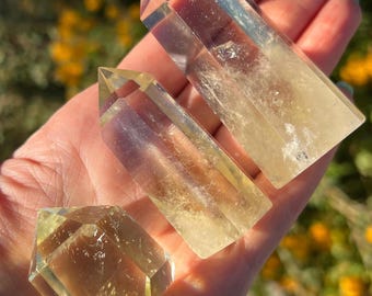 May include: Three yellow-toned crystal formations are held in a hand. The crystals have various shapes, including a diamond and two rectangular prisms with pointed tips. The crystals are translucent and appear to be polished.