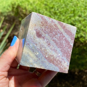 May include: A cube-shaped piece of polished stone with a mix of gray, white, and red colors. The stone has a swirling pattern and a smooth, polished surface.