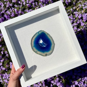 Puede incluir: Una rebanada de ágata enmarcada con bandas azules y blancas. La ágata se muestra sobre un fondo blanco y está enmarcada en un marco blanco.