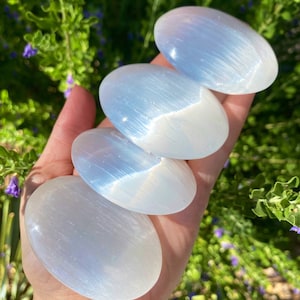 Puede incluir: Cuatro piedras de palma de selenita lisas y blancas con un brillo iridiscente y brillante. Las piedras están apiladas una encima de la otra, con la piedra más grande en la parte inferior y la más pequeña en la parte superior.