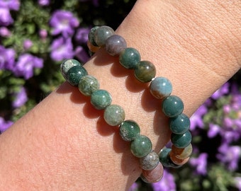 May include: Two bracelets made of green, brown, and white agate beads. The bracelets are stacked on a person's wrist.