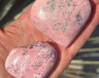 May include: Two heart-shaped stones with a pink and grey mottled pattern. The stones are smooth and polished, with a natural appearance. The hearts are held in a hand, showcasing their size and texture.