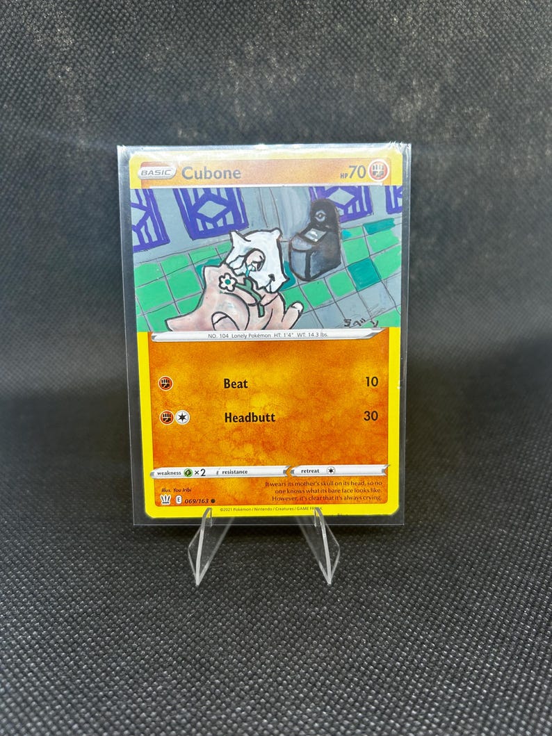 May include: A Pok&eacute;mon trading card featuring Cubone, with a yellow and orange design. The card shows an illustration of the Cubone character holding a bone. The card includes the text "Cubone", "Beat", and "Headbutt".