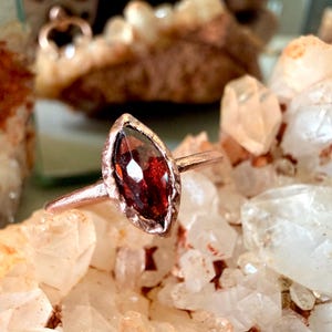 May include: A marquise-cut deep red garnet gemstone set in a textured copper band. The ring features a pointed, elongated stone with multiple facets that reflect light, held within a metallic setting that has a rustic, hammered appearance.
