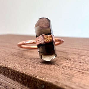 May include: A copper ring with a smoky quartz crystal set in the center. The ring is made of a thin band of copper with a textured finish. The crystal is a dark brown color with a slight shimmer.