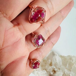 May include: Three copper wire-wrapped rings with red and pink gemstone settings. The rings vary in size, with the largest featuring a rectangular stone and the smallest a round stone. The gemstones have a mix of colors.