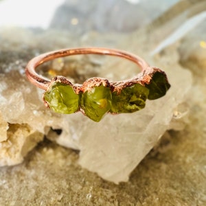 May include: A copper ring with four green peridot stones set in the band. The ring is on a white background.