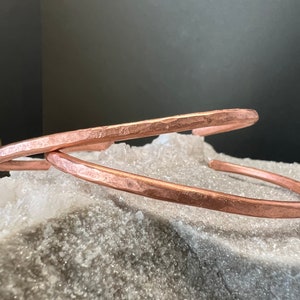 May include: Two hammered copper bracelets, one on top of the other, resting on a white, textured surface.