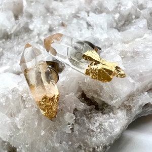 Könnte beinhalten: Ein Paar Ohrringe aus klarem Quarzkristall mit goldenen Blattverzierungen. Die Ohrringe sind auf einem weißen Hintergrund platziert.