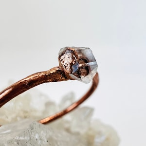 May include: A copper ring with a clear quartz crystal set on top. The ring is simple and delicate, with a raw, natural look.