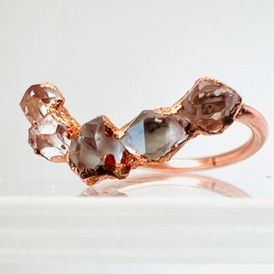 May include: A rose gold ring with a row of five raw, clear, and gray diamonds set in the band.