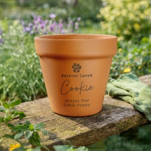 May include: A terracotta plant pot with the text "Forever Loved Cookie Always Our Little Puppy" and a paw print design. The pot is on a wooden surface, with a garden background of flowers and greenery.