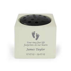 May include: A square, pale-coloured memorial flower vase with a black, circular, perforated top. The front of the vase features a pair of grey footprints and the inscription "Your tiny feet left footprints on our hearts" followed by a name and dates.