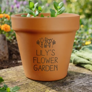 May include: A terracotta-coloured flower pot with the text "LILY'S FLOWER GARDEN" in black. The pot contains a small green plant. The pot has a decorative design of gardening tools. The pot is sitting on a wooden surface.