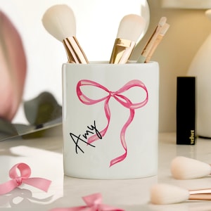 May include: White ceramic makeup brush holder featuring a watercolor pink bow and the name "Amy" in black script. The holder contains makeup brushes with gold handles and soft bristles.