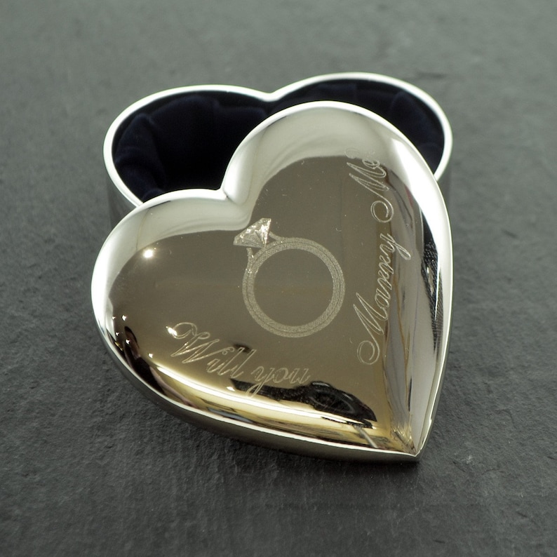 Op de afbeelding: Zilveren hartvormige ringdoos met een gegraveerde ring en de woorden "Will you marry me?"