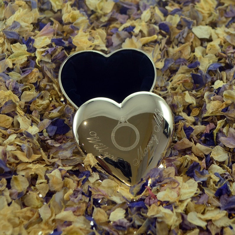 Op de afbeelding: Een zilveren hartvormige doos met een gegraveerde ring en de woorden "Will you marry me?" op een bed van gele en paarse bloemblaadjes.