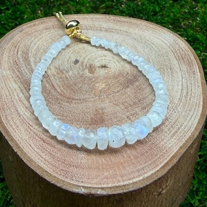 Op de afbeelding: Een armband bestaande uit kleine, gefacetteerde, doorschijnende witte maansteen kralen met een subtiele blauwe glans. De armband is voorzien van een goudkleurig metalen schuifsluiting waarmee de maat kan worden aangepast.
