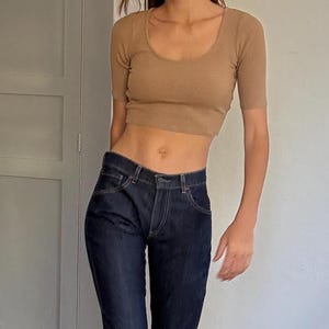 Op de afbeelding: Een damesoutfit met een lichtbruine crop top met korte mouwen en een ronde hals, gecombineerd met een donkerblauwe flared jeans. De outfit wordt gecompleteerd met witte sandalen met hakken. De setting is een kamer met houten vloeren en een witte muur.