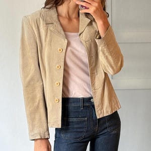 May include: A tan corduroy blazer with a button-up front is paired with a light pink top and dark wash denim jeans. The jeans have a flared leg. The outfit is completed with white sandals with a small heel.