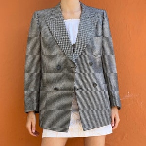 Può includere: Un blazer doppiopetto a quadri bianco e nero, abbinato a un top e una gonna bianchi. L'outfit è completato da stivali al ginocchio in camoscio marrone. L'ensemble è ambientato su uno sfondo arancione, mostrando uno stile di moda vintage.