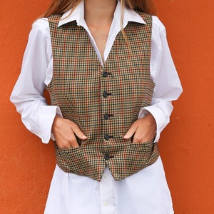 Op de afbeelding: Een persoon draagt een bruin en groen geruit vest over een wit overhemd, gecombineerd met lichtblauwe jeans en crèmekleurige laarzen. Het vest heeft een klassiek, getailleerd ontwerp met een knoopsluiting en zakken. De achtergrond is een effen oranje muur.