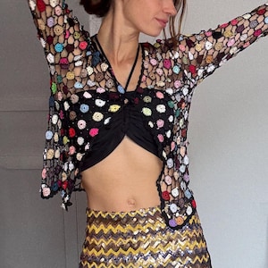 Può includere: Un top corto nero e una minigonna con paillettes con un motivo a zig-zag giallo e oro. Un top a maniche lunghe a trama aperta e colorata con vari cerchi colorati è indossato sopra il top corto. L'outfit è un insieme elegante.