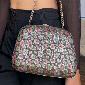 Op de afbeelding: Een rechthoekige clutch met een gouden kettingriem. De clutch is bedekt met een bloemenpatroon met rode, groene en witte bloemen op een zwarte achtergrond. De tas heeft een gouden sluiting.