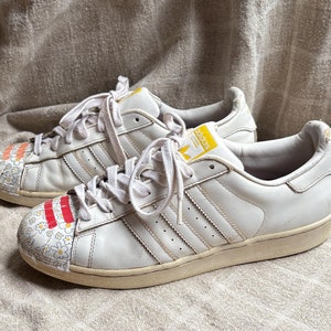 Puede incluir: Un par de zapatillas de cuero blanco con las tres rayas icónicas en los laterales. Las punteras presentan un diseño floral con detalles rojos y naranjas. La lengüeta tiene un logotipo amarillo.