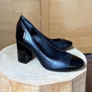 May include: A pair of black leather pumps with a glossy cap toe and a block heel. The shoes are displayed on a wooden tree stump, showcasing their design and texture. The heels are approximately 3 inches high.