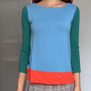 May include: A color-blocked long-sleeve top in blue, orange, and green, paired with a plaid pleated skirt. The outfit is completed with blue boots. The top has a boat neck and the skirt is mini length.