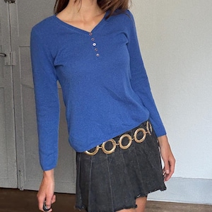 Puede incluir: Una camisa azul de manga larga con cuello en V y detalles de botones. Se combina con una falda plisada negra y un cinturón dorado. Una silla es visible en el fondo.