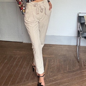May include: A woman wearing a black top with colorful polka dots and a sheer, open-weave overlay. She is wearing beige pants with a tie waist and black high-heeled sandals. The outfit is styled in a light-filled room with a wooden floor.