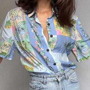 Puede incluir: Una camisa abotonada de manga corta con un diseño de patchwork en tonos azules, verdes, rosas y amarillos. La camisa presenta un estampado floral y paisajístico. El modelo lleva unos vaqueros de mezclilla de lavado claro.