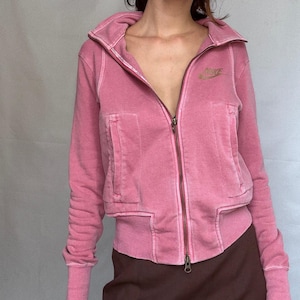 K&ouml;nnte beinhalten: Rosa Rei&szlig;verschlussjacke mit hohem Kragen und zwei Vordertaschen. Das Nike-Logo in Gold ist auf der Brust zu sehen. Der Rei&szlig;verschluss ist rosa. Die Jacke wird mit einem braunen Rock getragen.