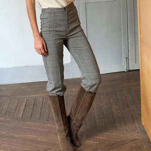 May include: A cream-colored, short-sleeved top with ruffled detailing paired with gray plaid trousers and brown suede knee-high boots. The outfit is captured in a mirror reflection, showcasing a casual yet stylish ensemble.